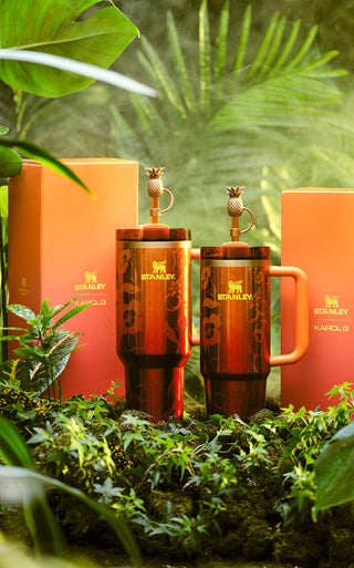 Stanley 1913 x Karol G copper-toned tumblers with pineapple lids and floral engravings, set in a lush tropical garden.