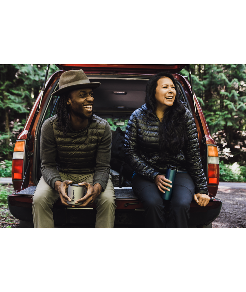 Man and woman sitting on the tailgate of a station wagon, each holding a Stanley titanium mug.