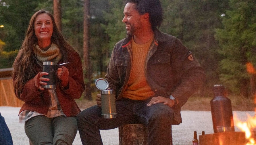 Woman and man, each holding a Stanley Bottle Opener Beer Stein, sitting next to a fire pit.