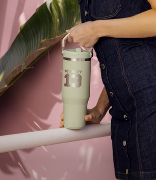 Woman in denim dress holding a IceFlow Tumbler in Lichen Green with 13 engraving, against a pink wall and tropical leaves