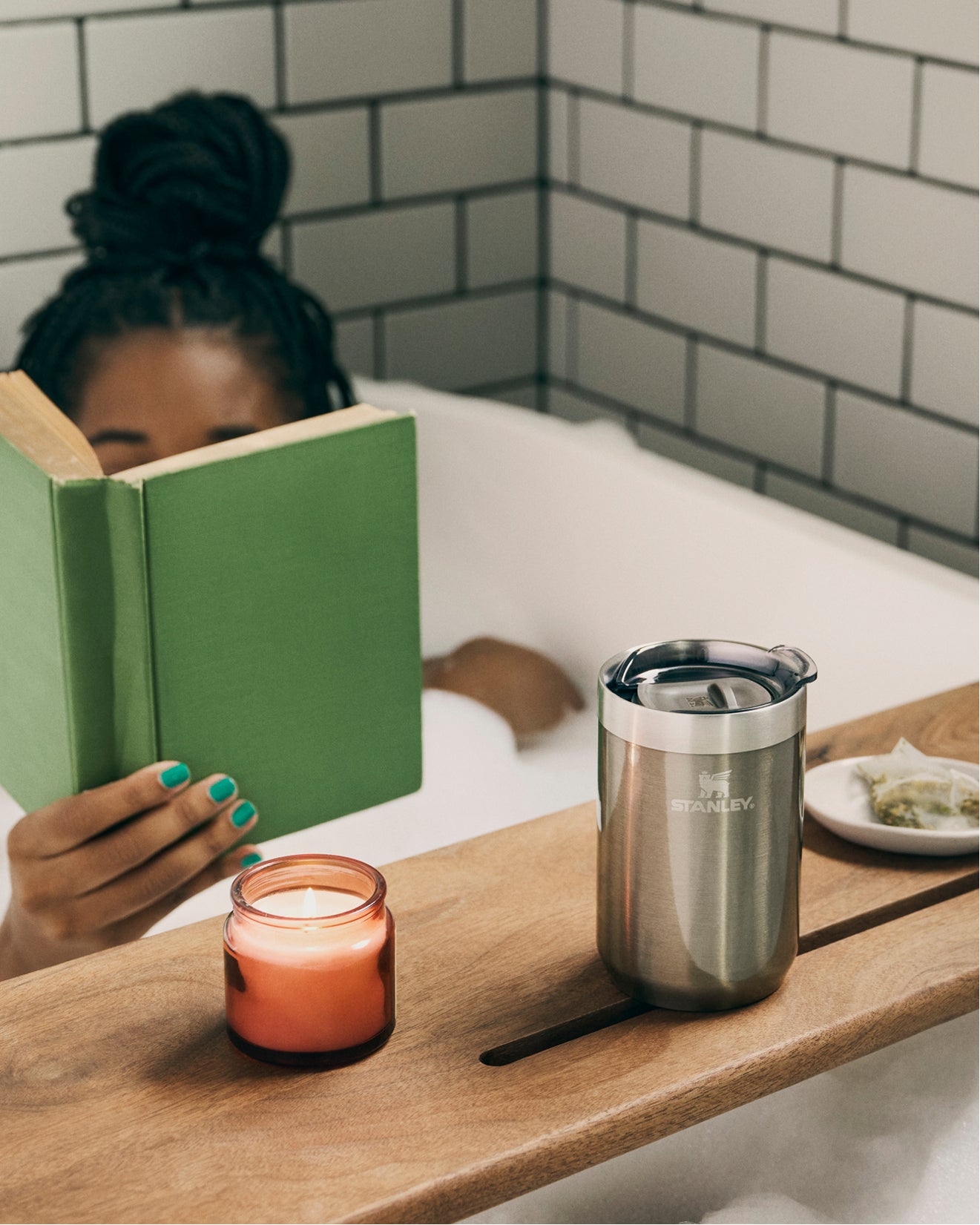 Person relaxing in a bubble bath reading a book with a lit candle and stainless steel Stanley mug on a wooden bath tray