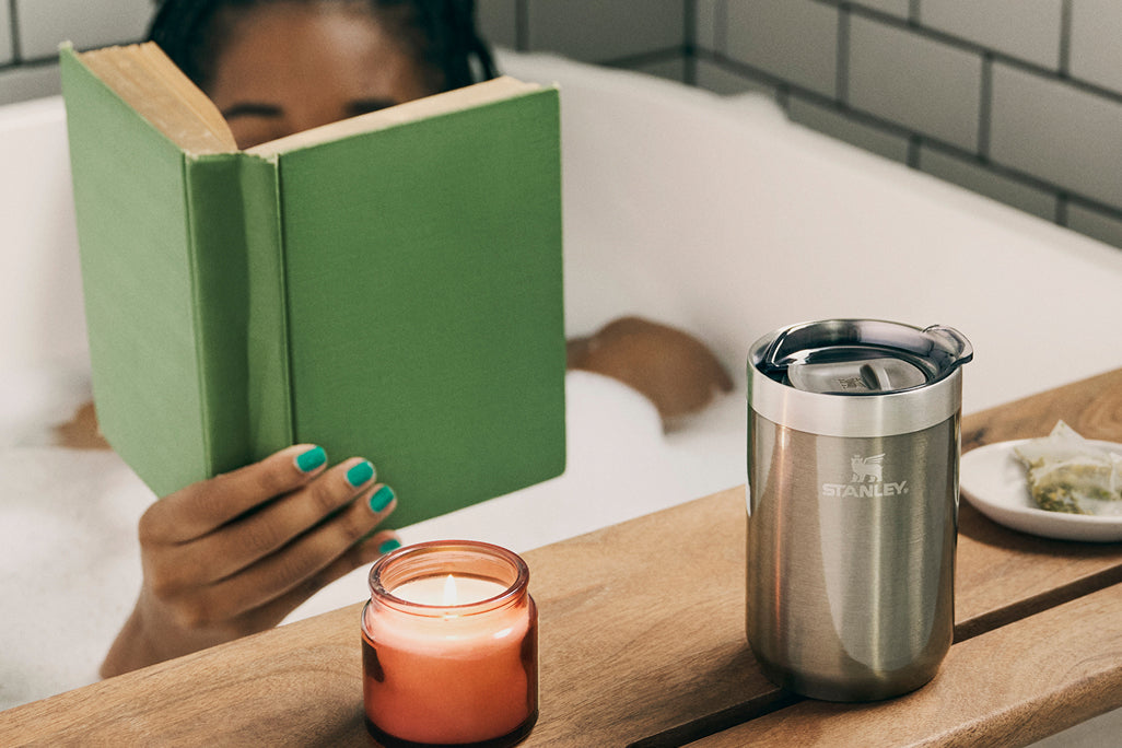 Person relaxing in a bubble bath reading a book with a lit candle and stainless steel Stanley mug on a wooden bath tray.