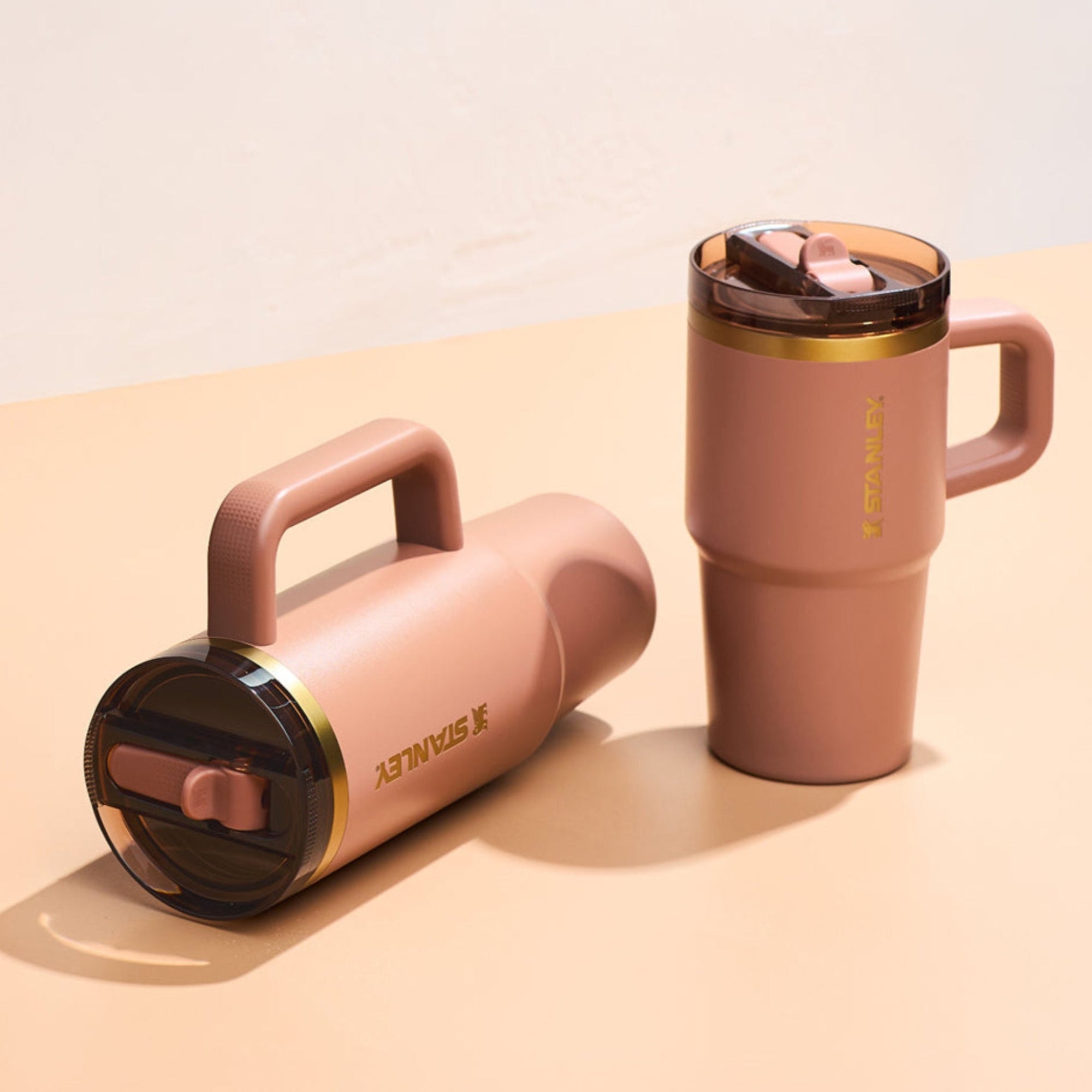 Blush pink Stanley travel mugs with gold accents, one upright and one on its side on a beige surface.