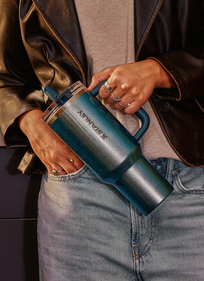 Person wearing jeans and a leather jacket holding a metallic blue Stanley tumbler with a handle and gradient finish.