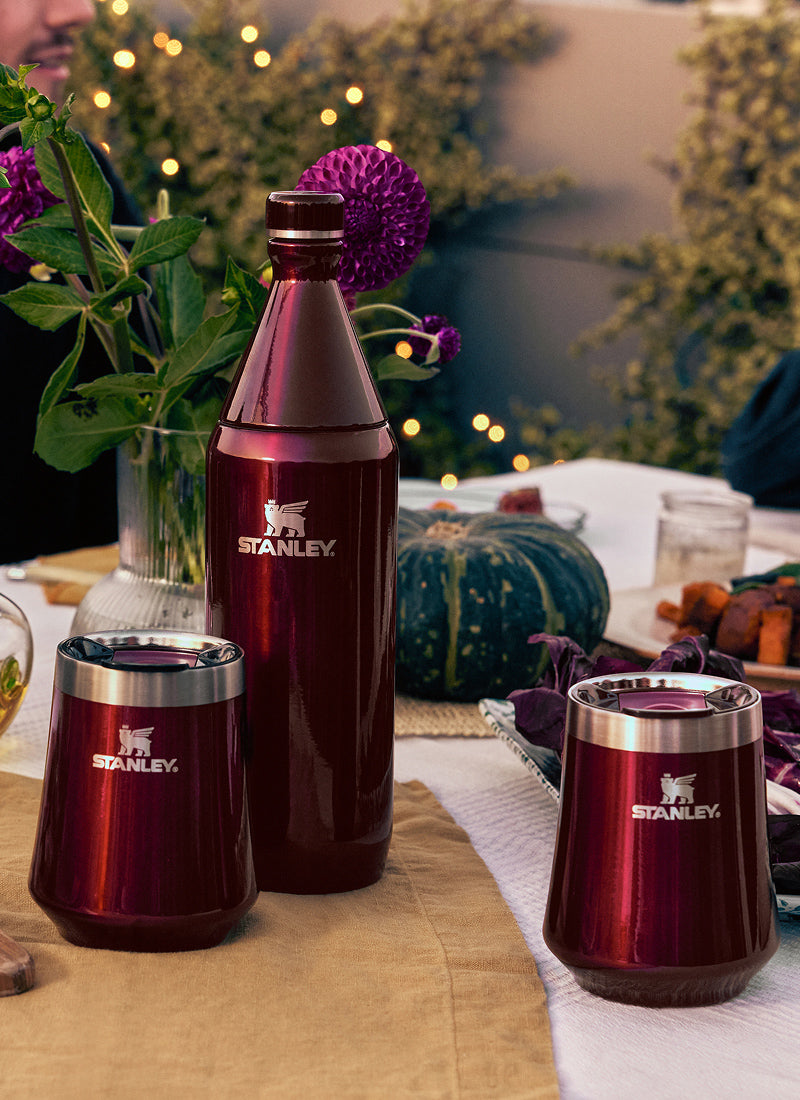 Three deep red Stanley drinkware pieces—a bottle and two tumblers—displayed on a dinner table with flowers and food in the background.
