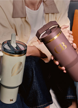 Two people holding Stanley tumblers with gold detailing—one sage green with a leaf design and one mauve with monogrammed initials “CB.”
