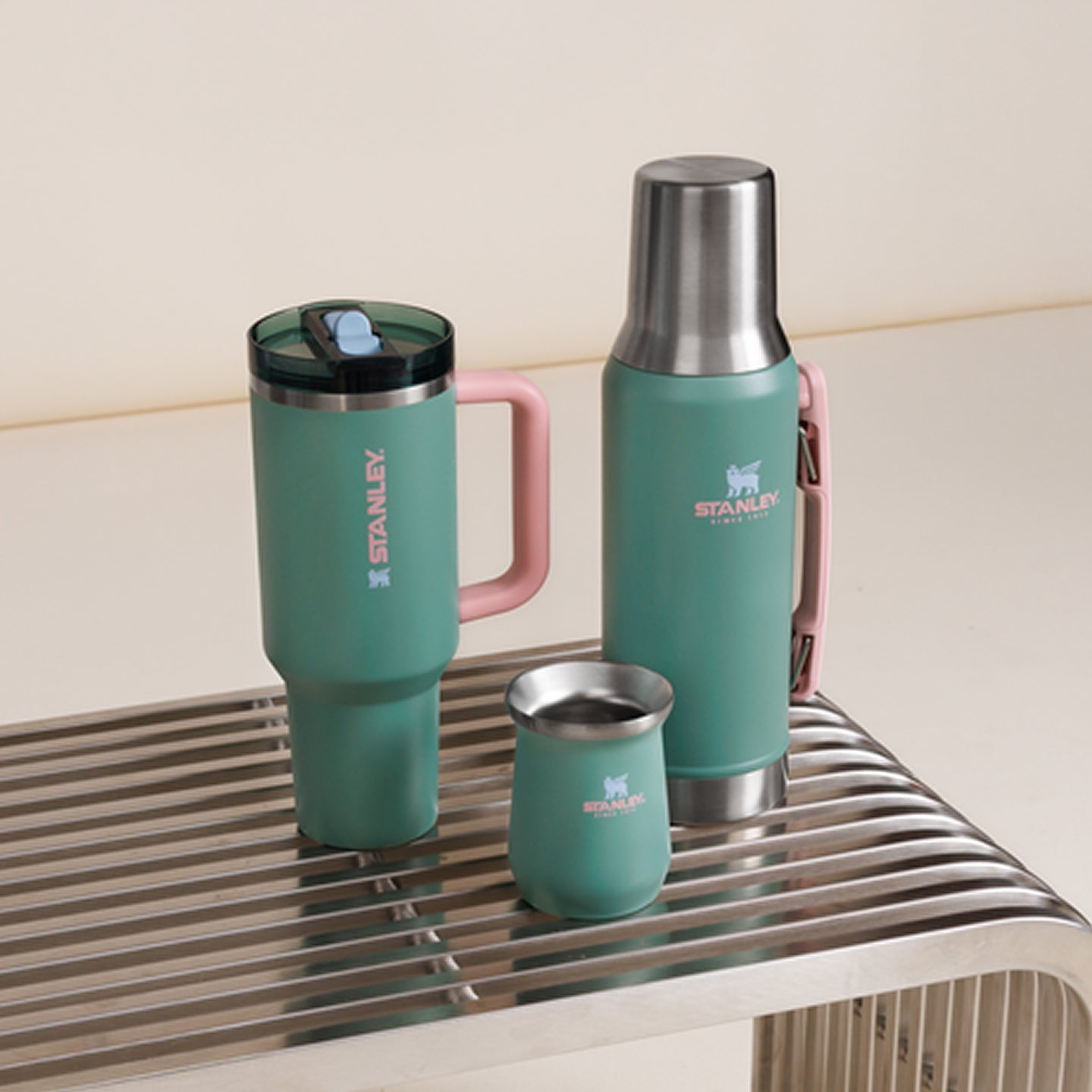 Three Stanley 1913 teal insulated drinkware pieces with pink handles displayed on a metal rack: tumbler, thermos, and small cup.
