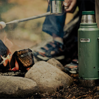 Stanley 1913 green Classic Vacuum Bottle beside a campfire as someone roasts marshmallows outdoors.
