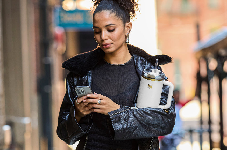 Woman walking outdoors holding a white Stanley 1913 tumbler with gold accents and straw while looking at her phone.