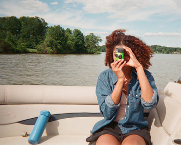 Person on a boat photographing a lake with a blue Stanley 1913 bottle resting on the seat beside her.
